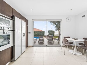 Een moderne keuken met een inbouwoven, een eetgedeelte met een ronde tafel en stoelen, en een schuifpui die uitkomt op een terras met buitenmeubels.