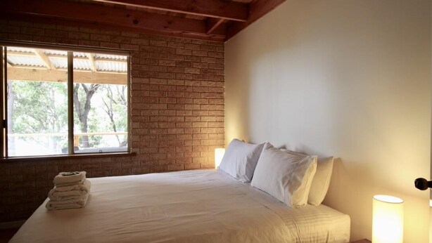 Quarto com cama, almofadas, abajur e janela com vista para árvores.