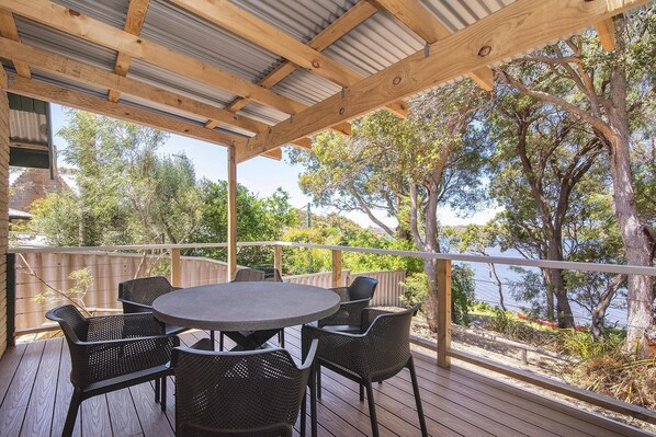 A covered outdoor area with a round table and chairs, surrounded by a wooden deck, overlooking a body of water and trees.