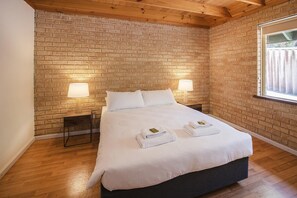 A hotel room with a brick wall, a wooden ceiling, a bed with white linens, two bedside lamps, and a window with curtains. - Riverscape Cottage No.11 - Riverscape - unit 11 with views over the Blackwood River and fire place (Augusta)