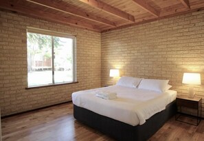 Un dormitorio con pared de ladrillo, vigas de madera en el techo, un ventanal grande, una cama con ropa blanca y dos lámparas de noche.