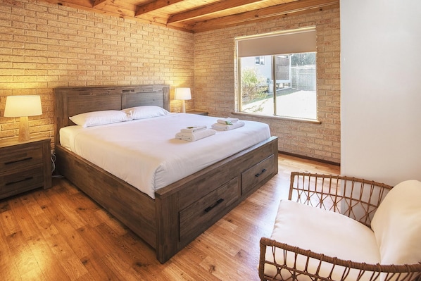Une chambre à coucher avec un lit en bois, deux tables de chevet avec des lampes, un fauteuil en osier et une grande fenêtre.