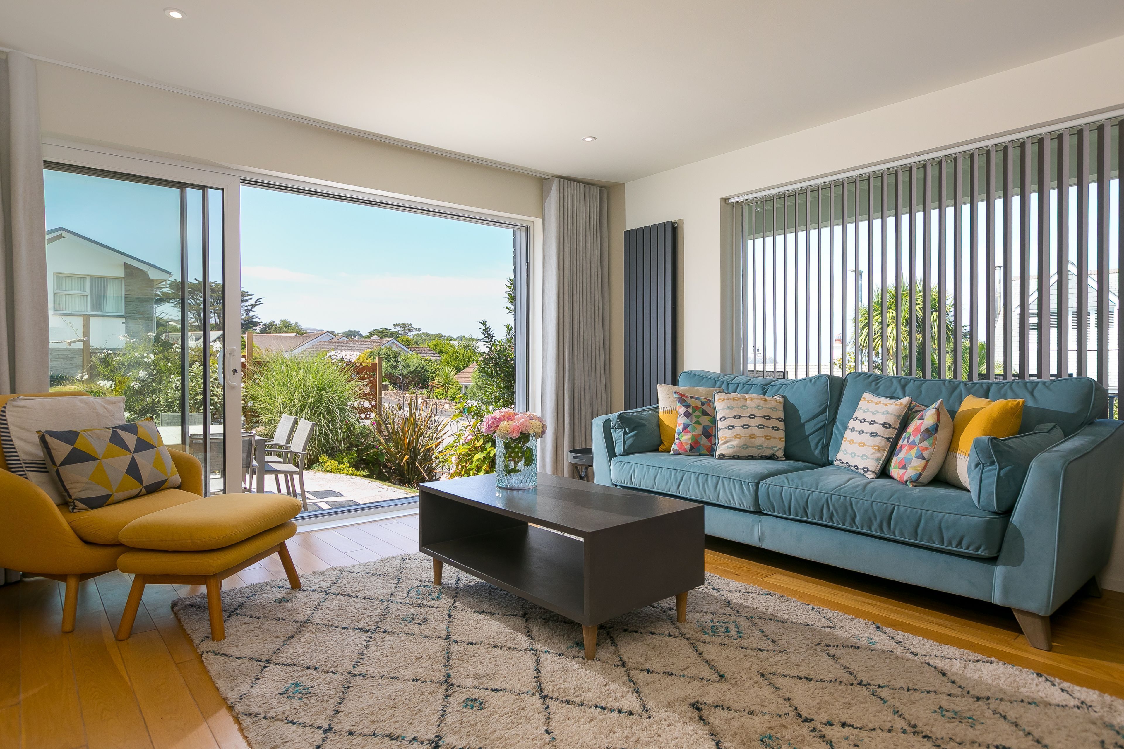 A modern living room with a sofa, armchair, and coffee table, large sliding glass doors, and a view of a residential area.