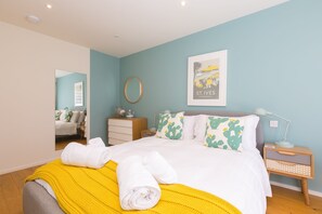 A neatly made bed with white bedding and a yellow throw, a bedside table with a lamp, and a framed picture on the wall. - Secret Beach, Carbis Bay – Sleeps 6 – Sea Views – Dog-Friendly – On-Site Parking (Carbis Bay)