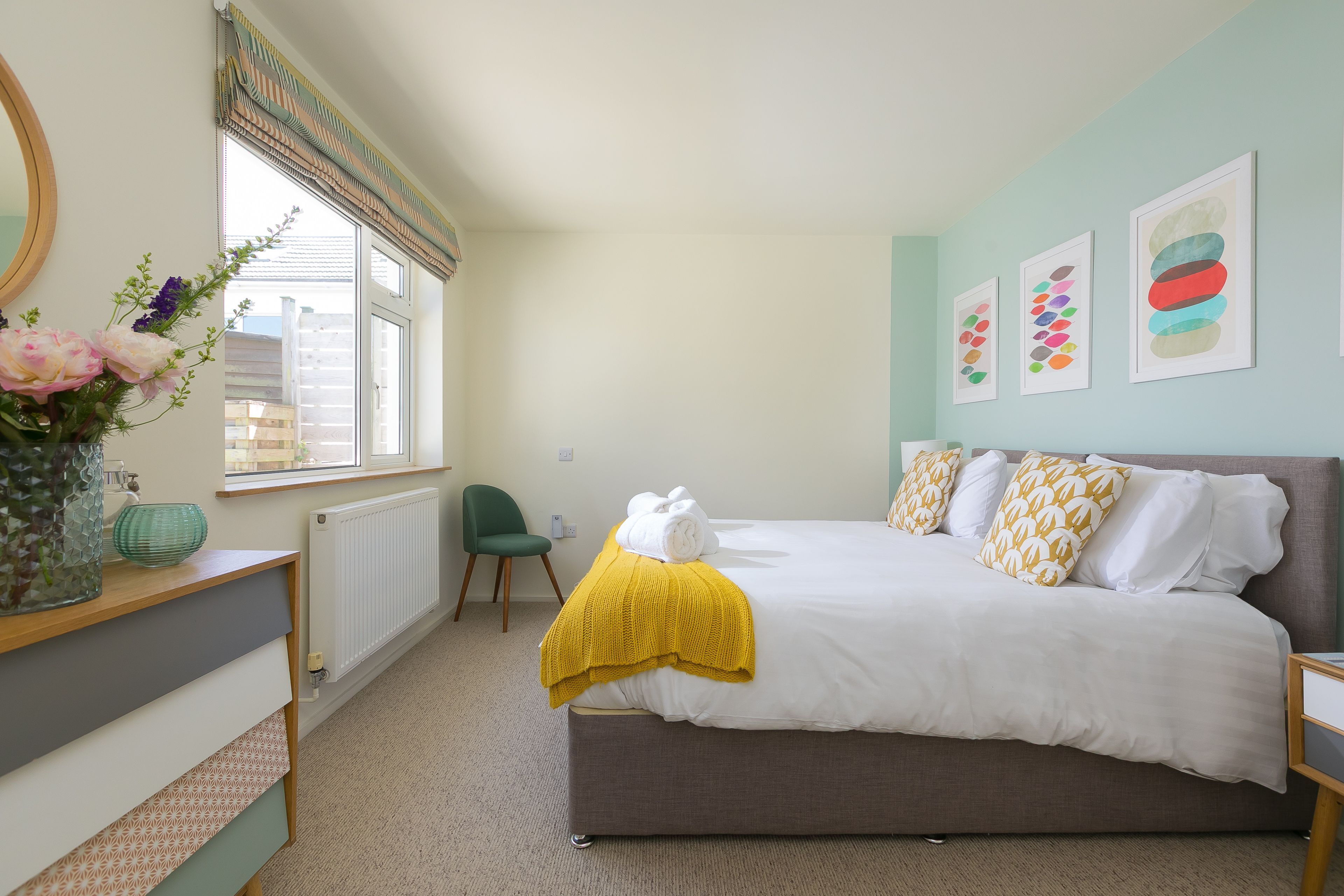 Une chambre à coucher avec un lit, une chaise, un radiateur, une fenêtre et des œuvres d’art accrochées au mur.