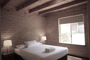 A bedroom with a brick wall, a large window, a bed with white linens, and two bedside lamps.