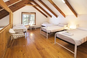 A room with wooden beams, two beds, a wicker chair, and a window with a view of trees.