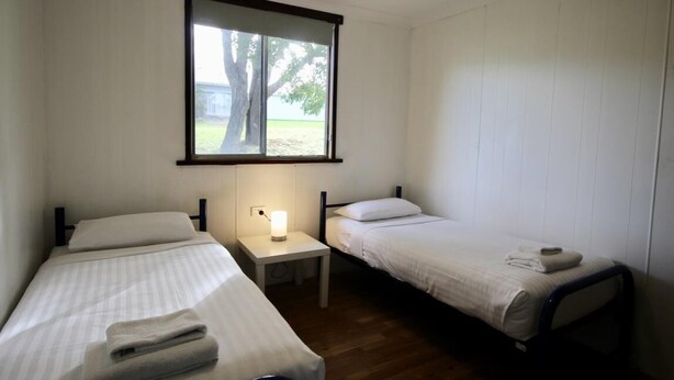 Una habitación pequeña y limpia con dos camas individuales, una mesita de noche con lámpara y ventana con vistas a árboles.