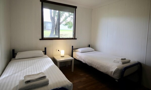 Una habitación pequeña y limpia con dos camas individuales, una mesita de noche con lámpara y ventana con vistas a árboles.