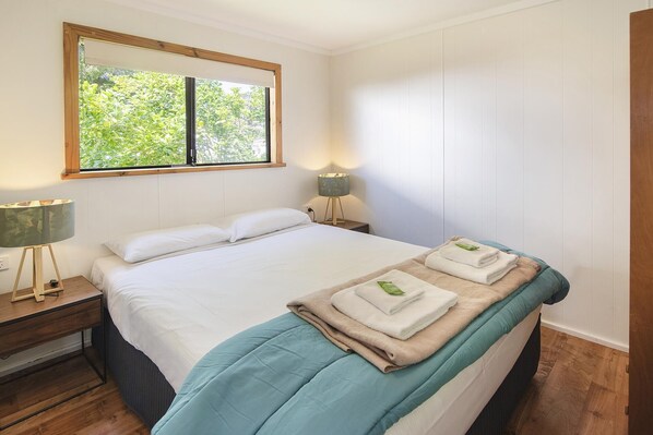 A bedroom with a bed, bedside lamps, a window with a view of greenery, and a wooden floor.