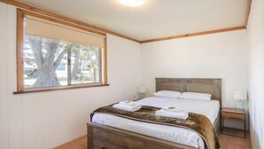 Un dormitorio con cabecera de madera, un ventanal grande, una cama con ropa blanca, una mesita de noche con lámpara y un aparador de madera con cajones.