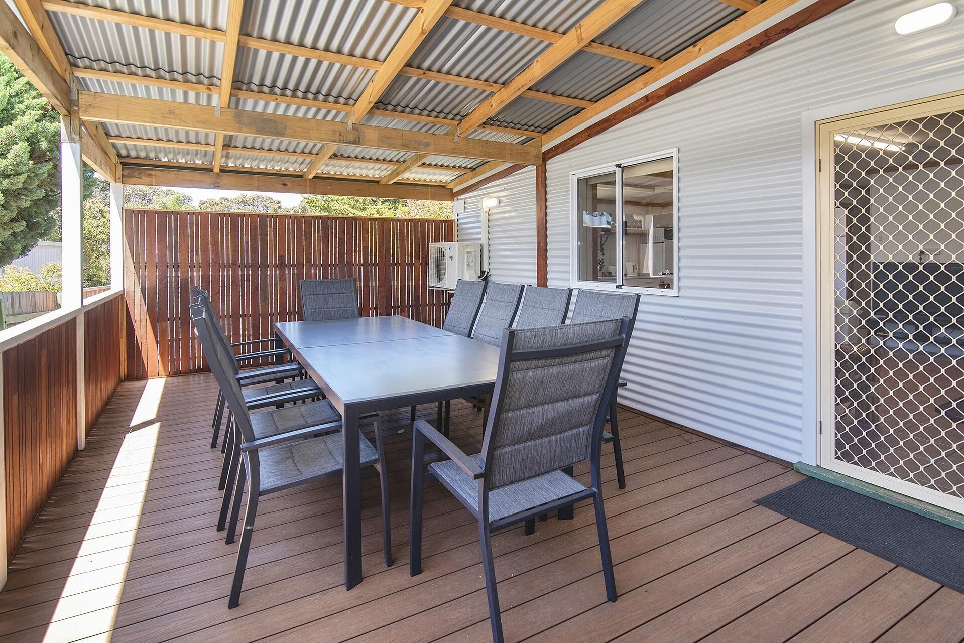 Un patio techado al aire libre con una mesa y sillas de comedor, una terraza de madera y una cerca metálica.