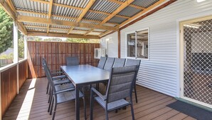 Un patio techado al aire libre con una mesa y sillas de comedor, una terraza de madera y una cerca metálica.