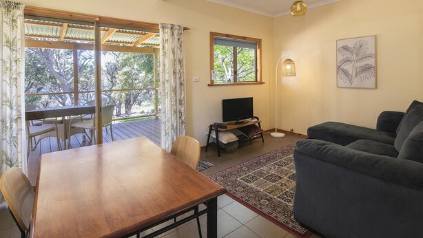 A living room with a wooden table, a sofa, a TV, and a window with a view of trees. - Riverscape Cottage No.8 with River Views - Riverscape - unit 8 with views over the Blackwood River (Augusta)