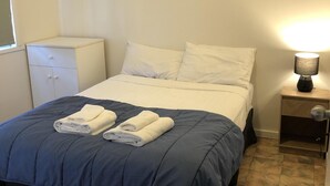Een net opgemaakt bed met een blauwe deken en twee opgevouwen witte handdoeken, een bedlampje en een klein wit nachtkastje met een lade.