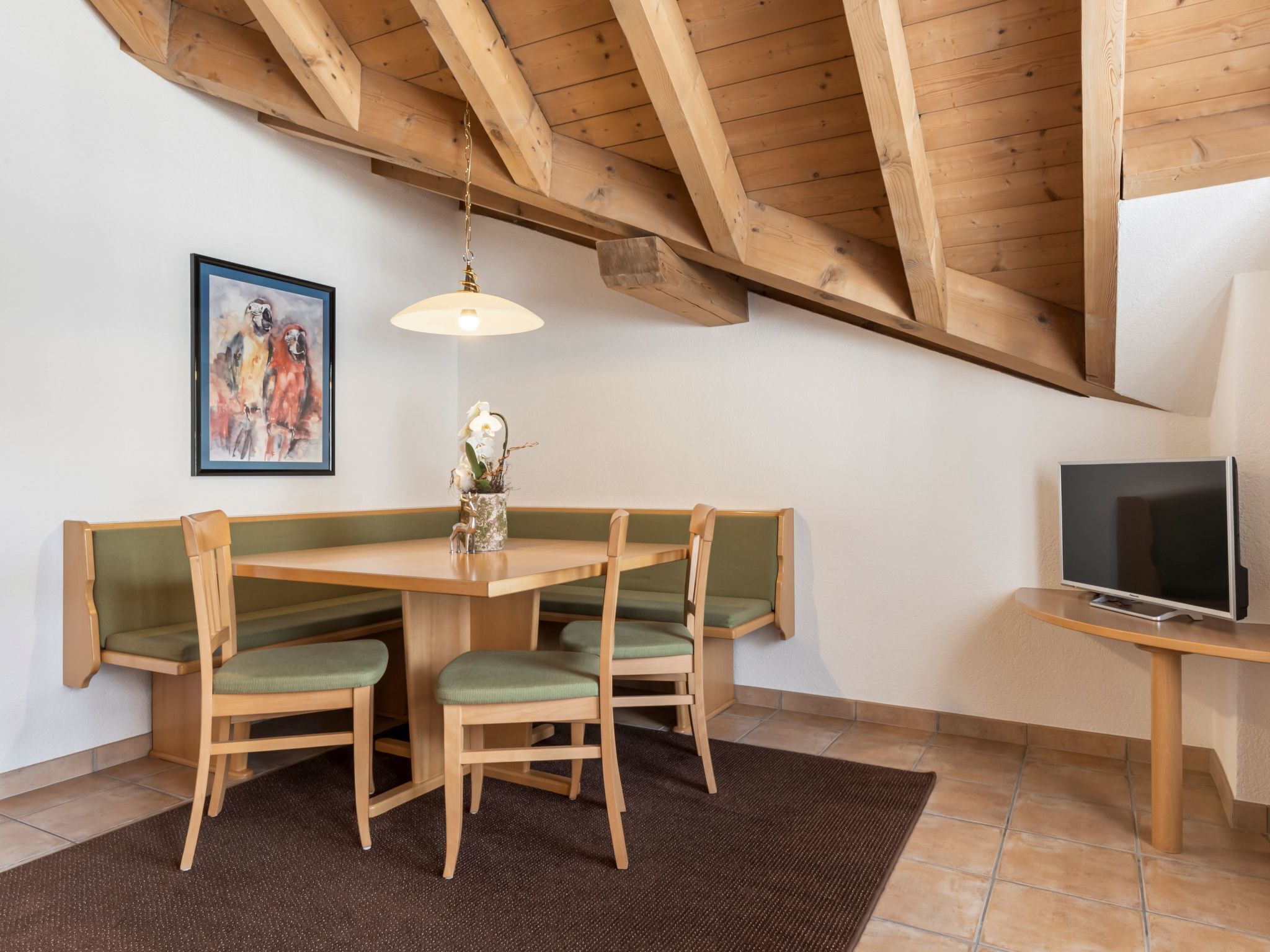 A dining area with a wooden table and chairs, a painting on the wall, a TV on a stand, and a ceiling with exposed beams.