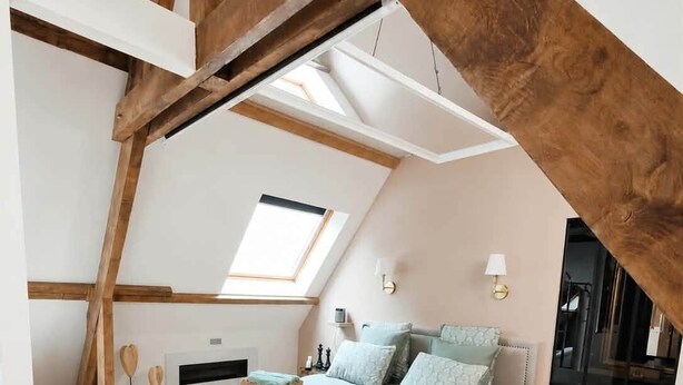 Un dormitorio con techo inclinado, tragaluz, cama con ropa de cama azul claro, mesita de noche y una silla.