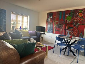 A living room with a brown leather sofa, a glass coffee table, a TV, and a dining area with blue chairs. - Super Comfy, Convenient two Bedroom Apartment (Corsham)