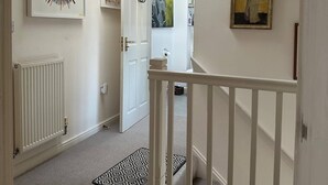 A hallway with a white door, a radiator, a black and white patterned rug, a ceiling light, and a painting on the wall. - Super Comfy, Convenient two Bedroom Apartment (Corsham)