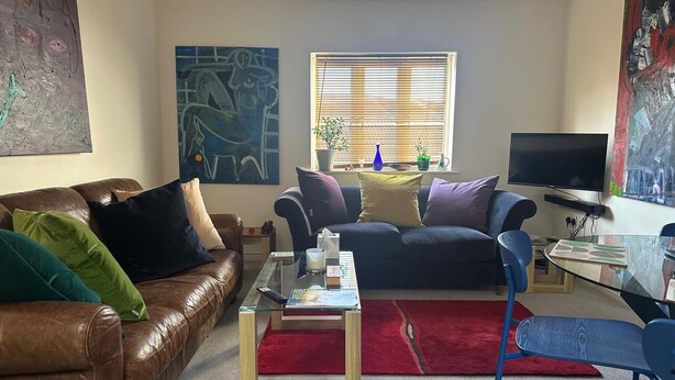 A living room with a brown leather sofa, a glass coffee table, a red rug, a television, and abstract wall art. - Super Comfy, Convenient two Bedroom Apartment (Corsham)