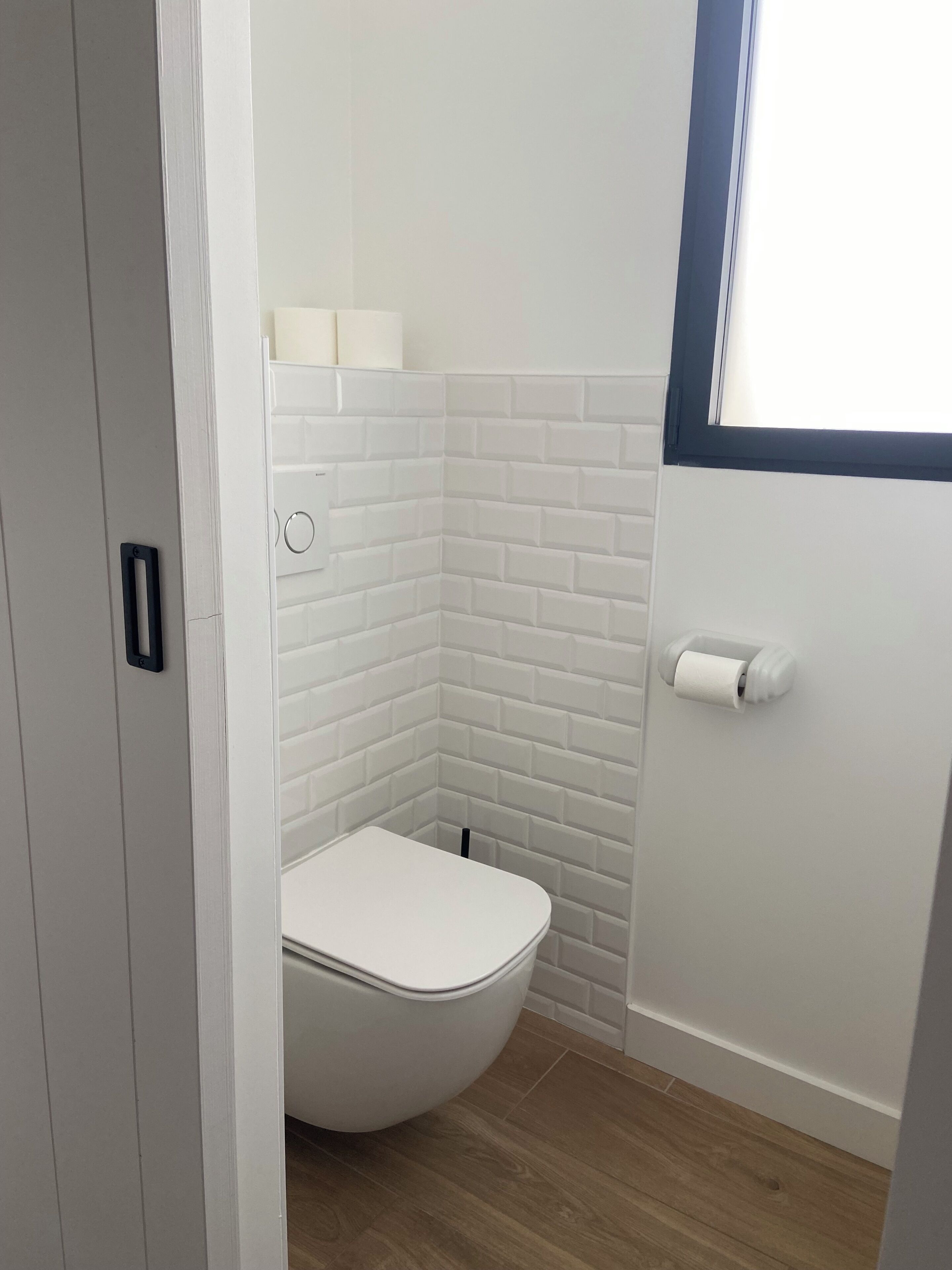 Une petite salle de bain moderne avec des toilettes blanches, des carreaux de céramique blancs de type « métro » et un plancher en bois.