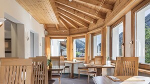Interior de madera con mesas y sillas, ventanas amplias y vistas a montañas.
