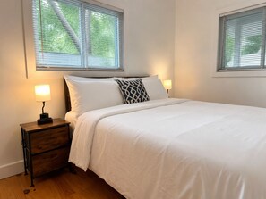 A neatly made bed with white linens, two bedside lamps, a wooden nightstand, and a window with blinds. - Lakefront Cabin | 3BR | Centrally Located (Perry)