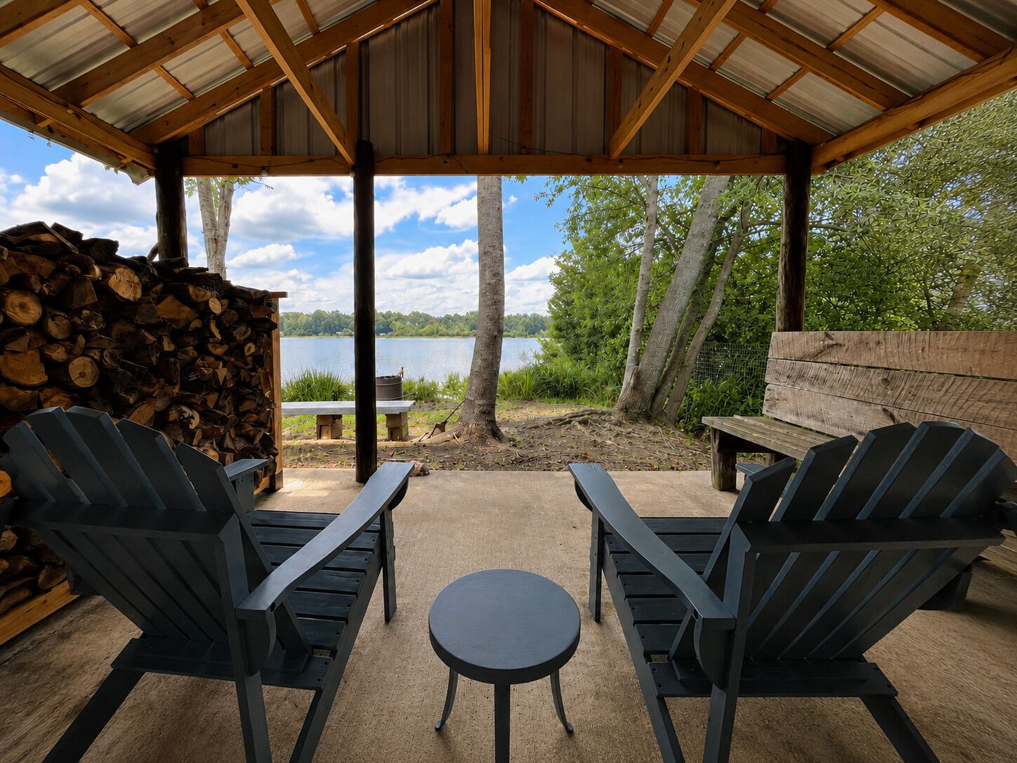 An outdoor seating area with Adirondack chairs and a small round table, overlooking a lake.