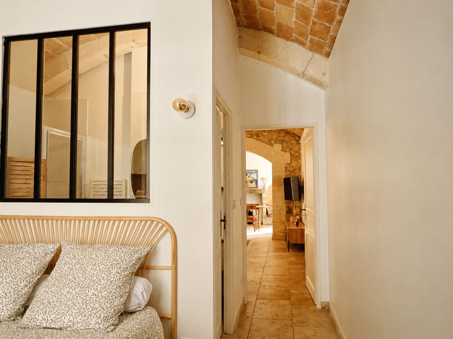 A room with a bed, a mirror, and a door leading to a hallway with a stone wall and a television.