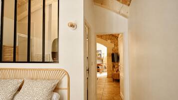 A room with a bed, a mirror, and a door leading to a hallway with a stone wall and a television.