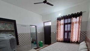 A bedroom with a bed, a ceiling fan, a window with curtains, a door, and a radiator. - Sehrawat Guest House (Sakrai)