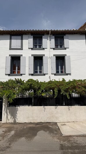 Um edifício de dois andares com paredes brancas e persianas cinzentas, base de concreto e cerca de metal preto.