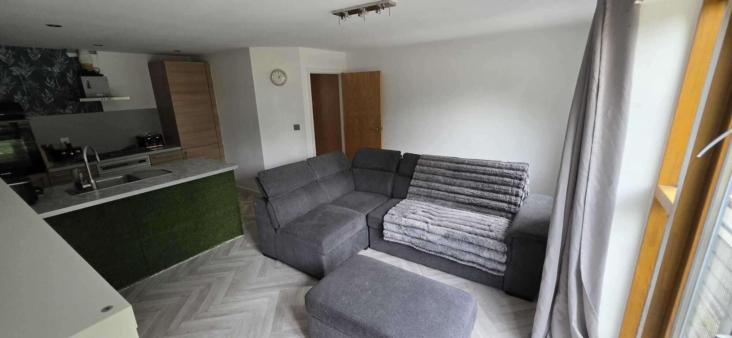 A modern living room with a grey sectional sofa, a small kitchen area, and a herringbone-patterned floor.