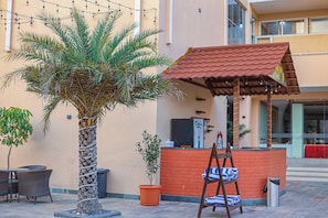 Una palmera, asientos al aire libre, una pequeña construcción con techo rojo y un muro de ladrillo.