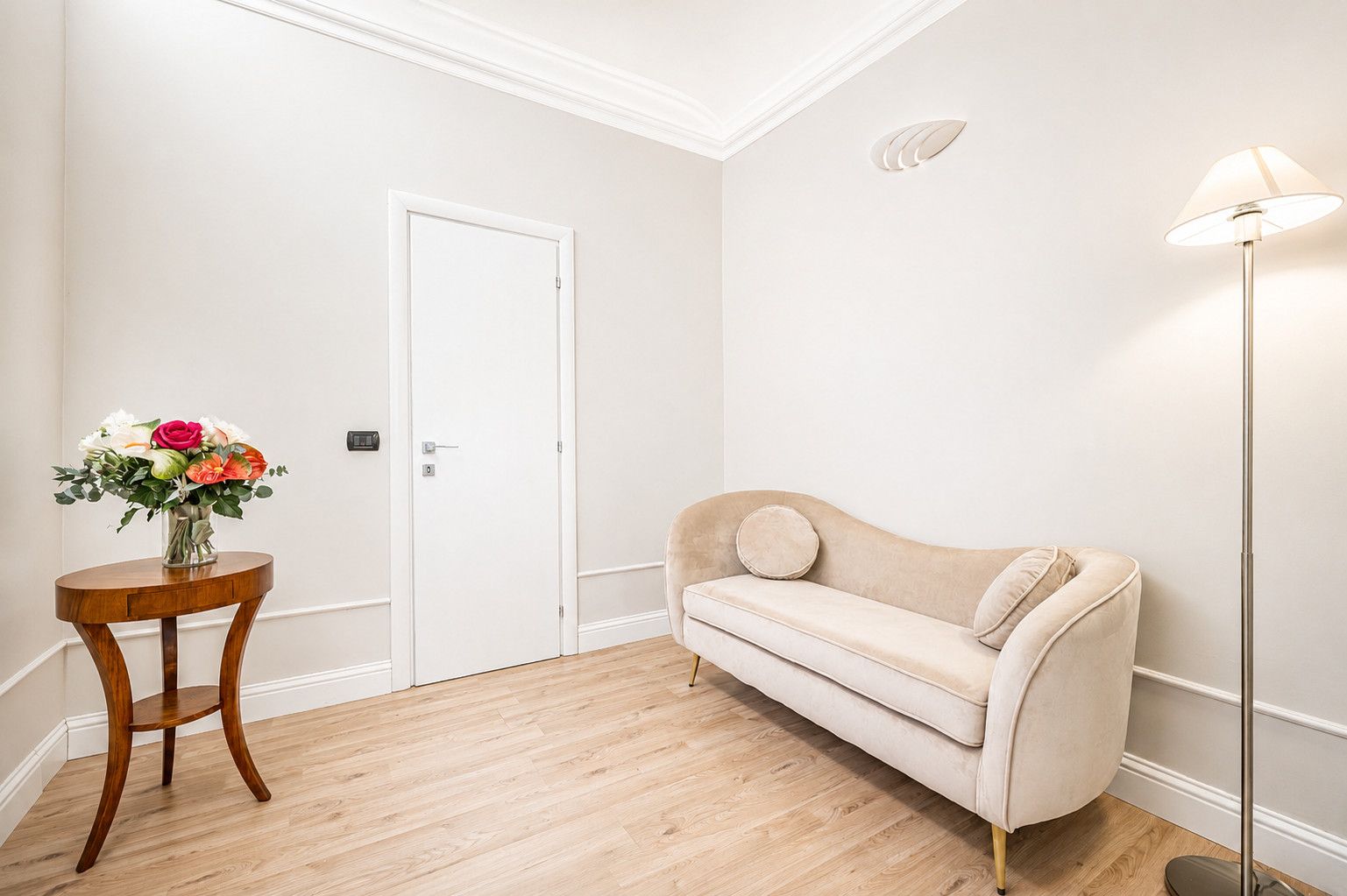 Une pièce avec un canapé beige, une table ronde en bois sur laquelle est posé un vase de fleurs, une porte blanche et un lampadaire.