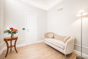 A room with a beige sofa, a round wooden table with a vase of flowers, a white door, and a floor lamp. - HB Room 1 (Florence)
