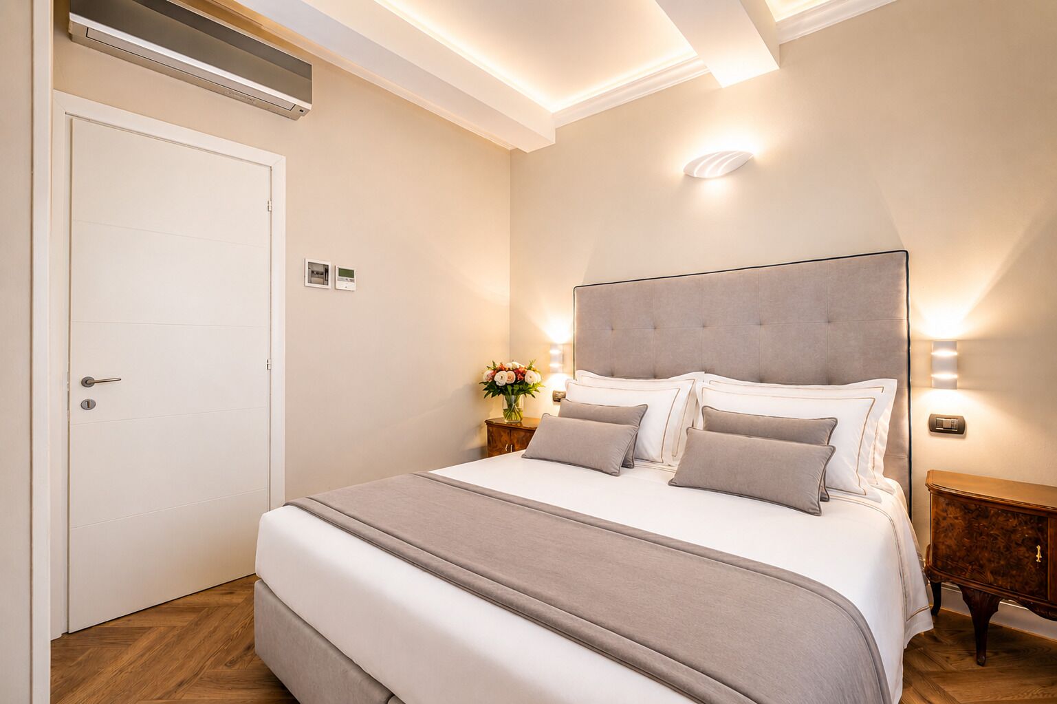 A bedroom with a large bed, a bedside table with a vase of flowers, a wall-mounted light, and a door with a lock.