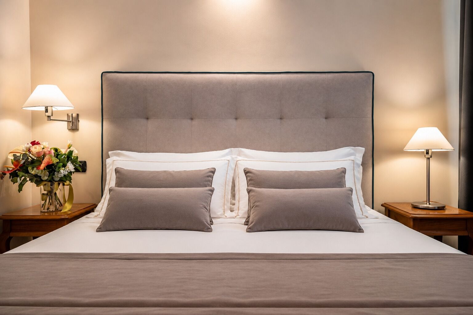 Een net opgemaakt bed met een gestoffeerd hoofdbord, twee nachtkastjes met lampen, en een vaas met bloemen aan de zijkant.