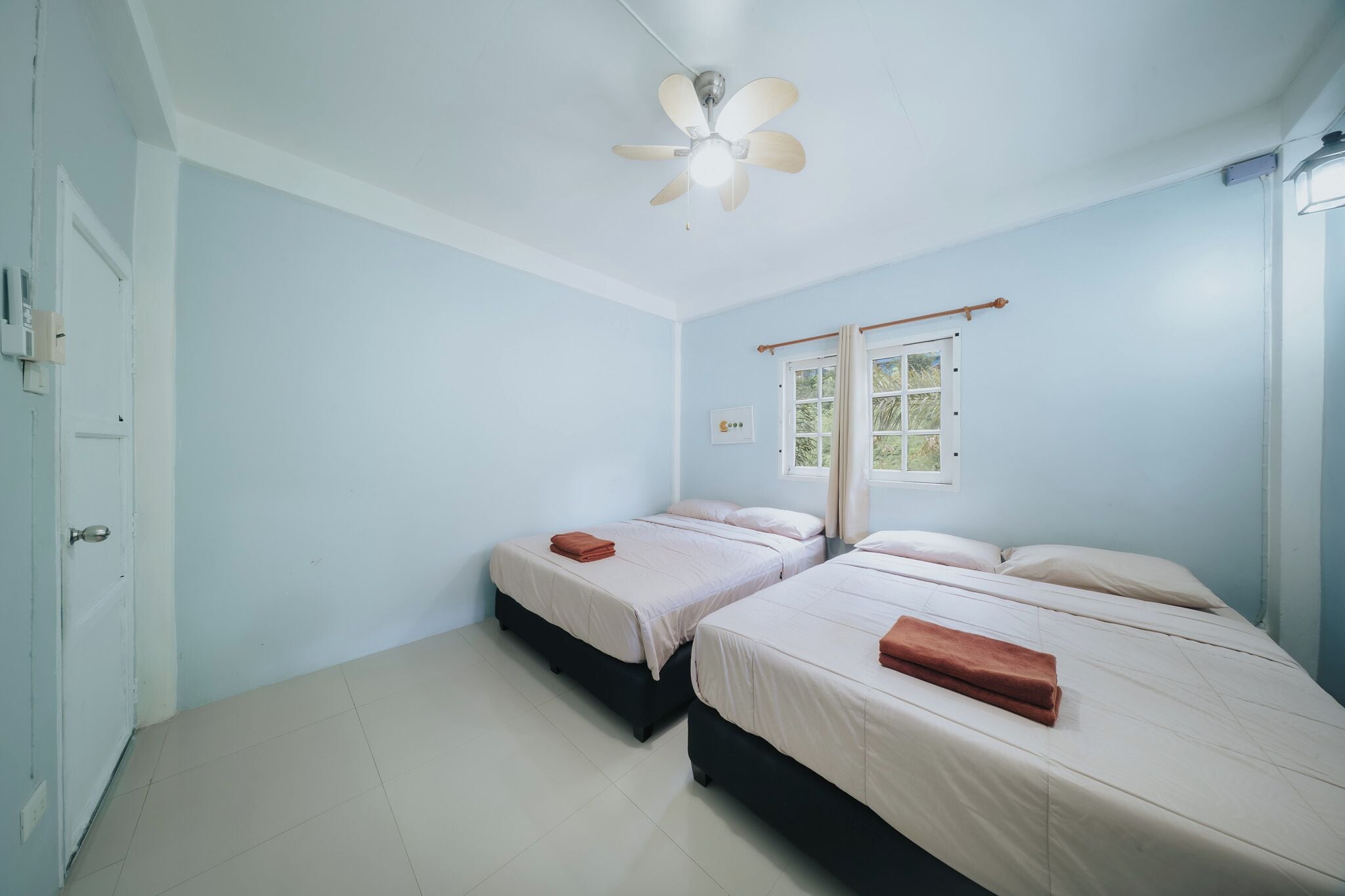 A room with two beds, a ceiling fan, a window with curtains, and a door.