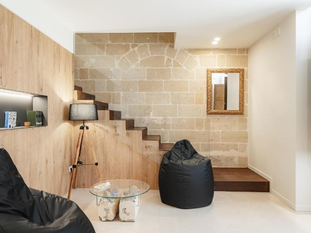 A modern living room with a stone wall, a wooden staircase, a black bean bag, and a glass coffee table.