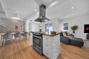 A modern kitchen with a central island, a dining area with chairs, a sofa, and a TV. - Villa Maris, Modern 5BR Getaway Near Miami Beach (North Miami)