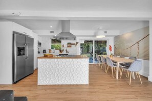 A modern kitchen with a central island, a dining area with a wooden table and chairs, and a view of the outdoors. - Villa Maris, Modern 5BR Getaway Near Miami Beach (North Miami)