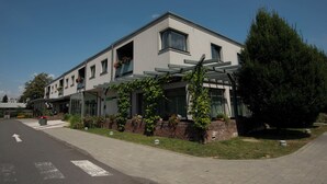 A modern two-story building with a flat roof, large windows, and a covered outdoor area with plants.