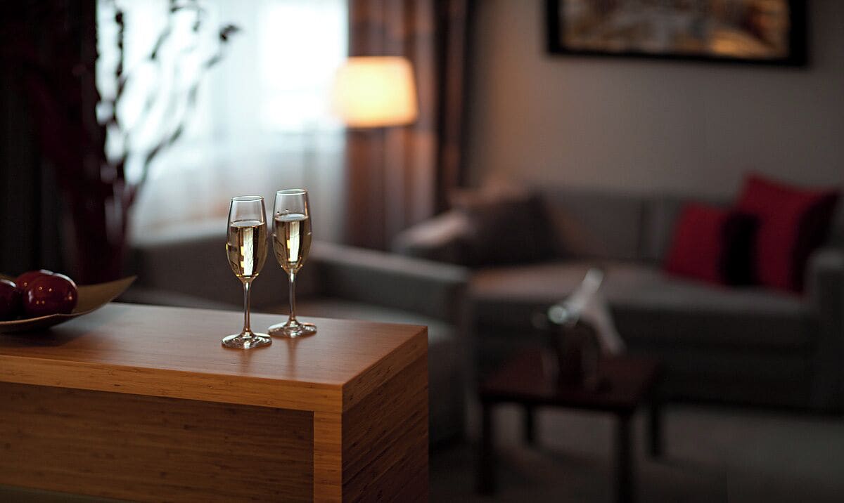 A wooden table with two glasses of a clear liquid, possibly champagne, in a room with a couch and a lamp in the background.