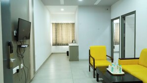 A hotel room with a television, a desk, two chairs, a sofa, and a window with curtains. - PLA Nest -Comfort Stay (Thanjavur)