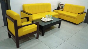 A living room with yellow sofas and wooden chairs, a coffee table with a tray of bottles and cups, and a door with a handle. - PLA Nest -Comfort Stay (Thanjavur)