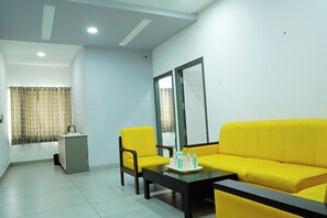 A room with a yellow sofa, a small table with a tray of bottled water, a chair, a window with curtains, and a small kitchenette area. - PLA Nest -Comfort Stay  (Thanjavur)