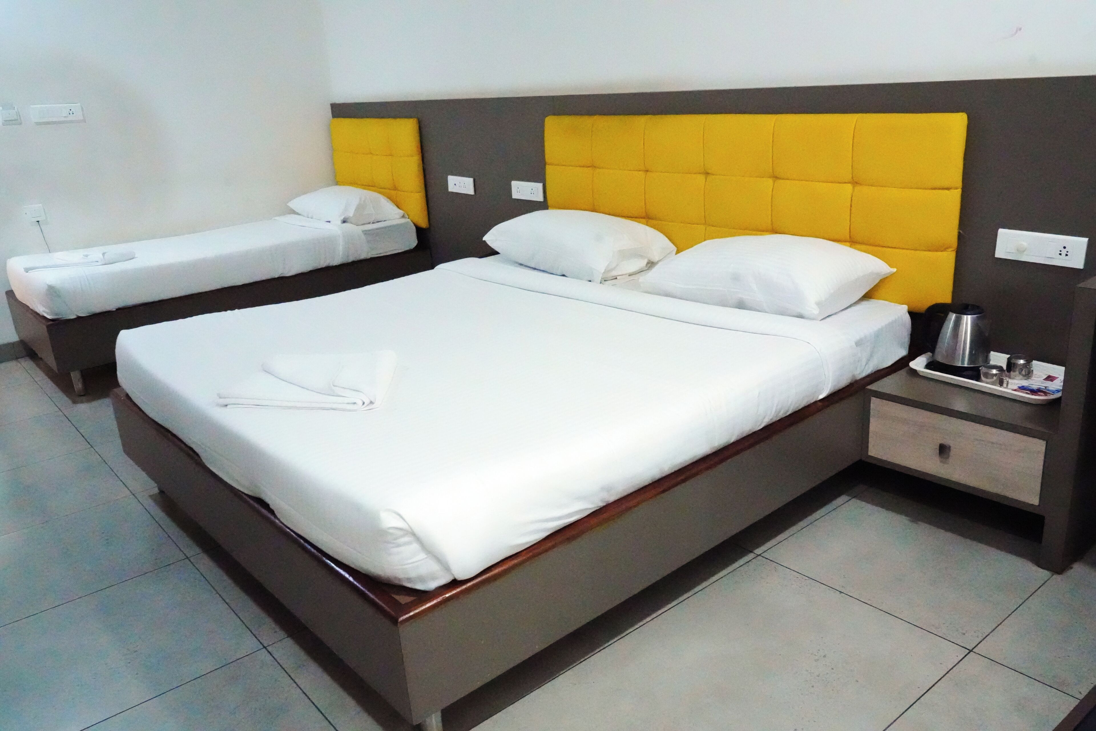 Una habitación de hotel con una cama grande, una cama más pequeña, una mesita de noche con hervidor y un enchufe de pared.
