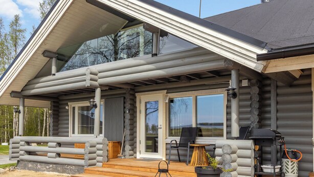 A log cabin with a covered porch, a grill, and a large window.