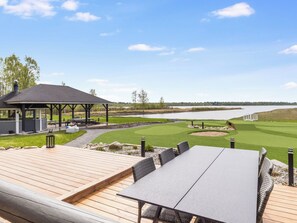 Ett trädäck med ett bord och stolar, en överbyggd paviljong och utsikt över en golfbana och en sjö.